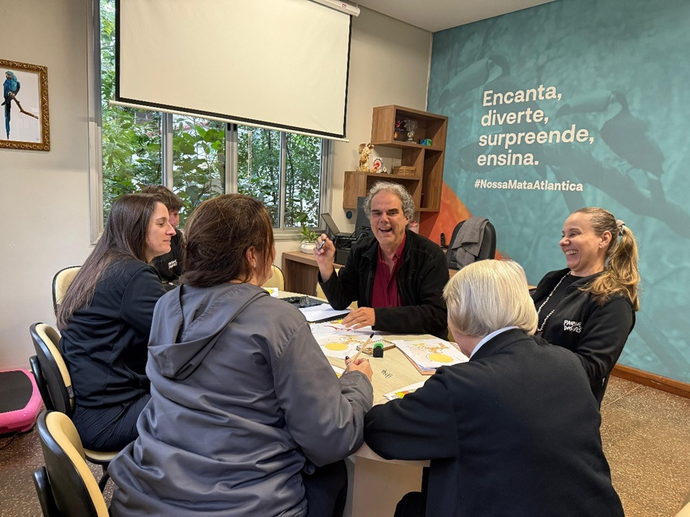 Cinco pessoas sentadas ao redor de uma mesa, em ambiente interno, participando de uma atividade em grupo. Ao fundo, há uma parede azul com a frase "Encanta, diverte, surpreende, ensina. #NossaMataAtlantica".