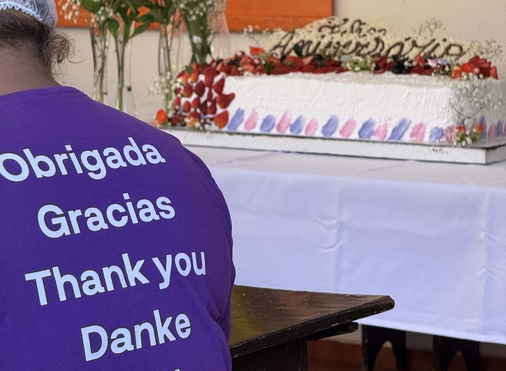 Camiseta com a palavra “obrigada” escrita em vários idiomas e, ao fundo, um bolo decorado com frutas e flores.