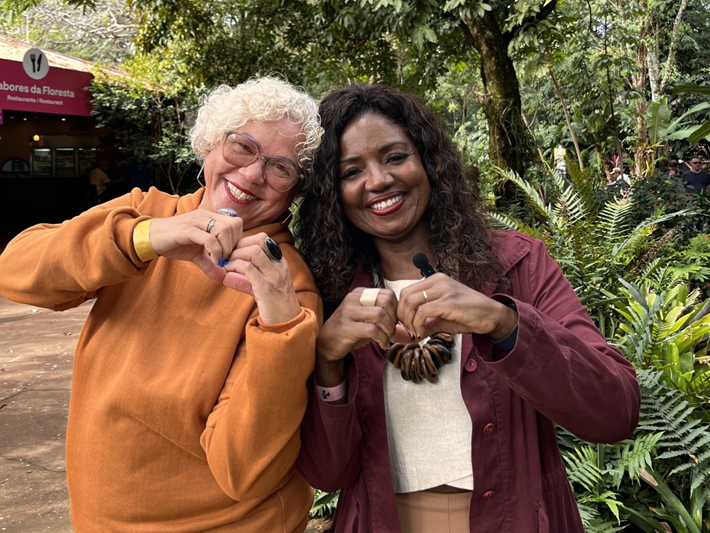 Duas mulheres di Caldeirão com Mion sorrindo fazem o gesto de coração com as mãos em frente ao Restaurante Sabores da Floresta, no Parque das Aves.