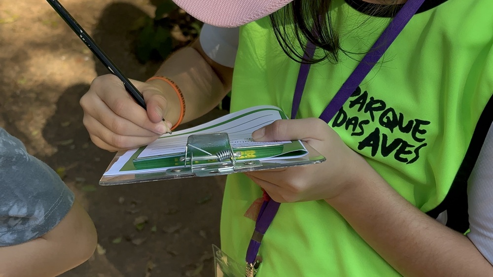 Criança com colete do Parque das Aves anota observações em uma prancheta durante a atividade.