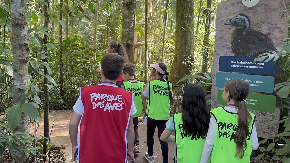 Crianças com coletes coloridos do Parque das Aves caminham pela trilha acompanhadas por educadora ambiental.