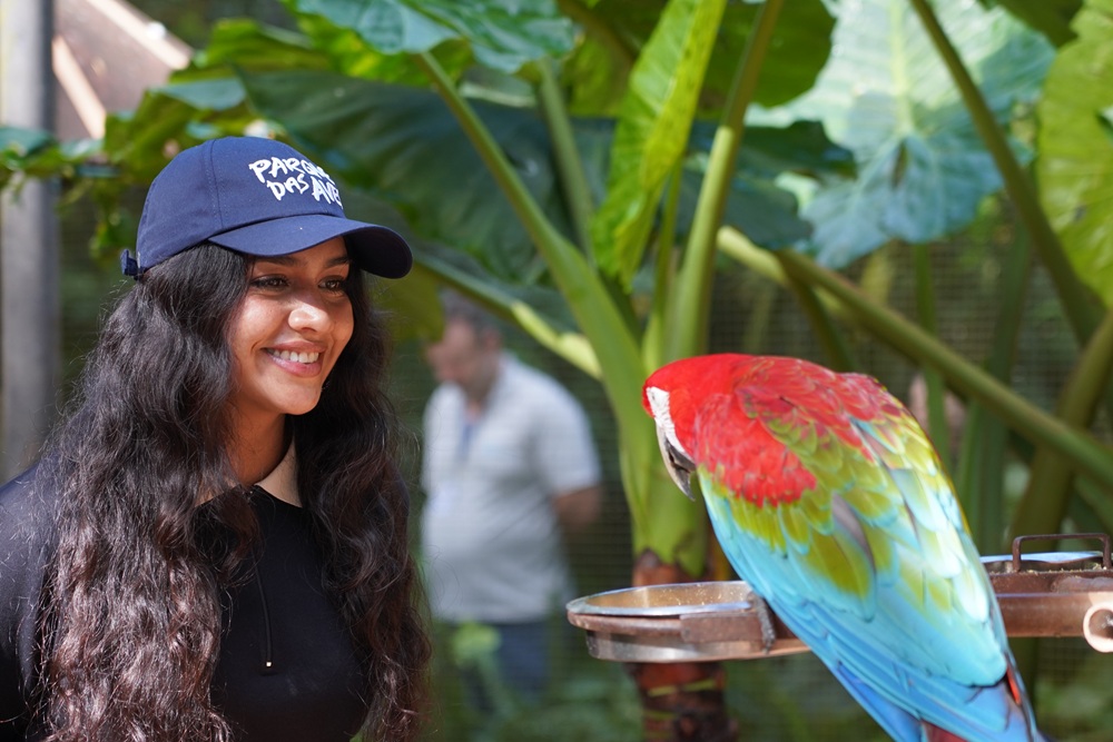 Mulher com boné do Parque das Aves observa uma arara-vermelha sobre o poleiro.